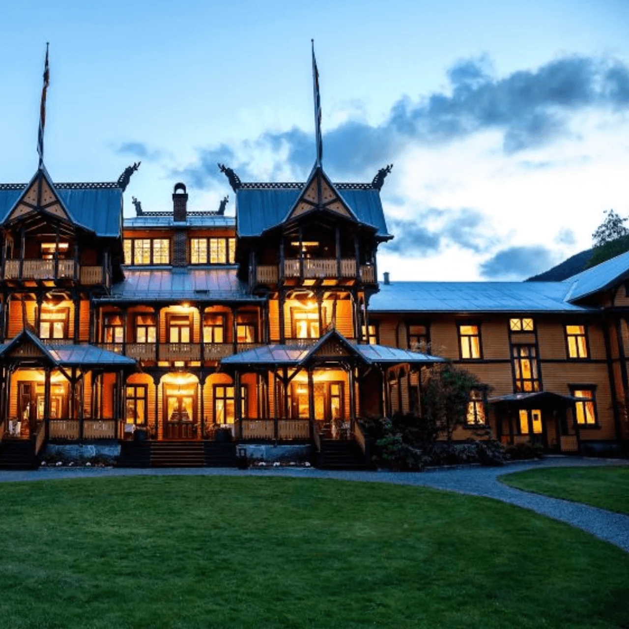 An ornate, illuminated wooden building with towers stands against a twilight sky, set amid grassy lawns and mountains in the background.