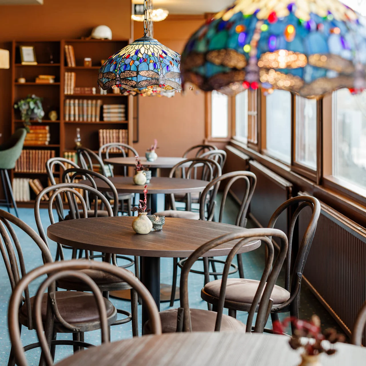 Runda träbord omges av stolar; färgglada lampor hänger ovanför. Bokhyllor med dekorationer står mot en brun vägg i ett varmt belyst kafé med stora fönster.