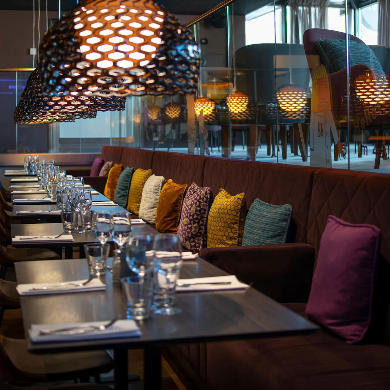 En rad middagsbord är dukade med glas och servetter, kompletterade med färgglada kuddar på en brun soffa i en stilig, stämningsfull restaurang.