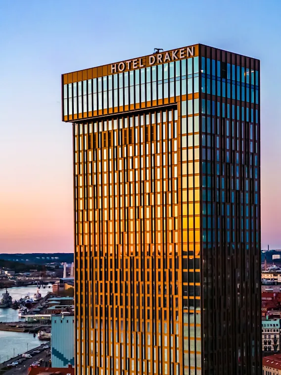 Ett högt, modernt hotell med texten "HOTEL DRAKEN" reflekterar solljus vid en flod i solnedgången, omgivet av stadslandskap inklusive byggnader och en bro.