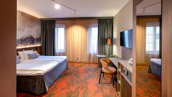 A well-furnished hotel room features a large bed, wall art, and a desk area with chairs. It is brightly lit with floor-to-ceiling curtains and a wall-mounted TV.
