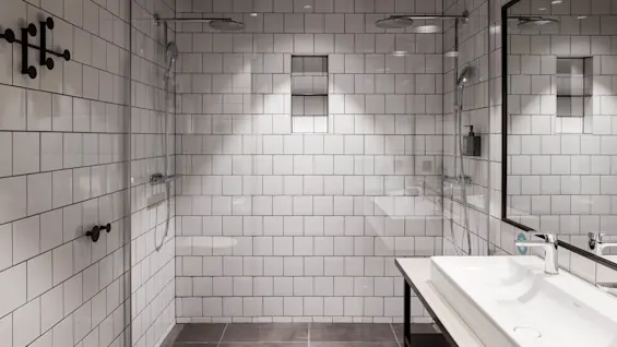 A modern bathroom with two showerheads, white tiled walls, and a large mirror reflecting a sink. The space is clean and well-lit, creating a sleek, minimalist design.