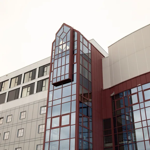 Moderni arkkitehtoninen rakennus, jossa on suuri, monikerroksinen lasiosa, joka ulottuu ulospäin, täydennettynä punaisilla ja harmailla paneeleilla.