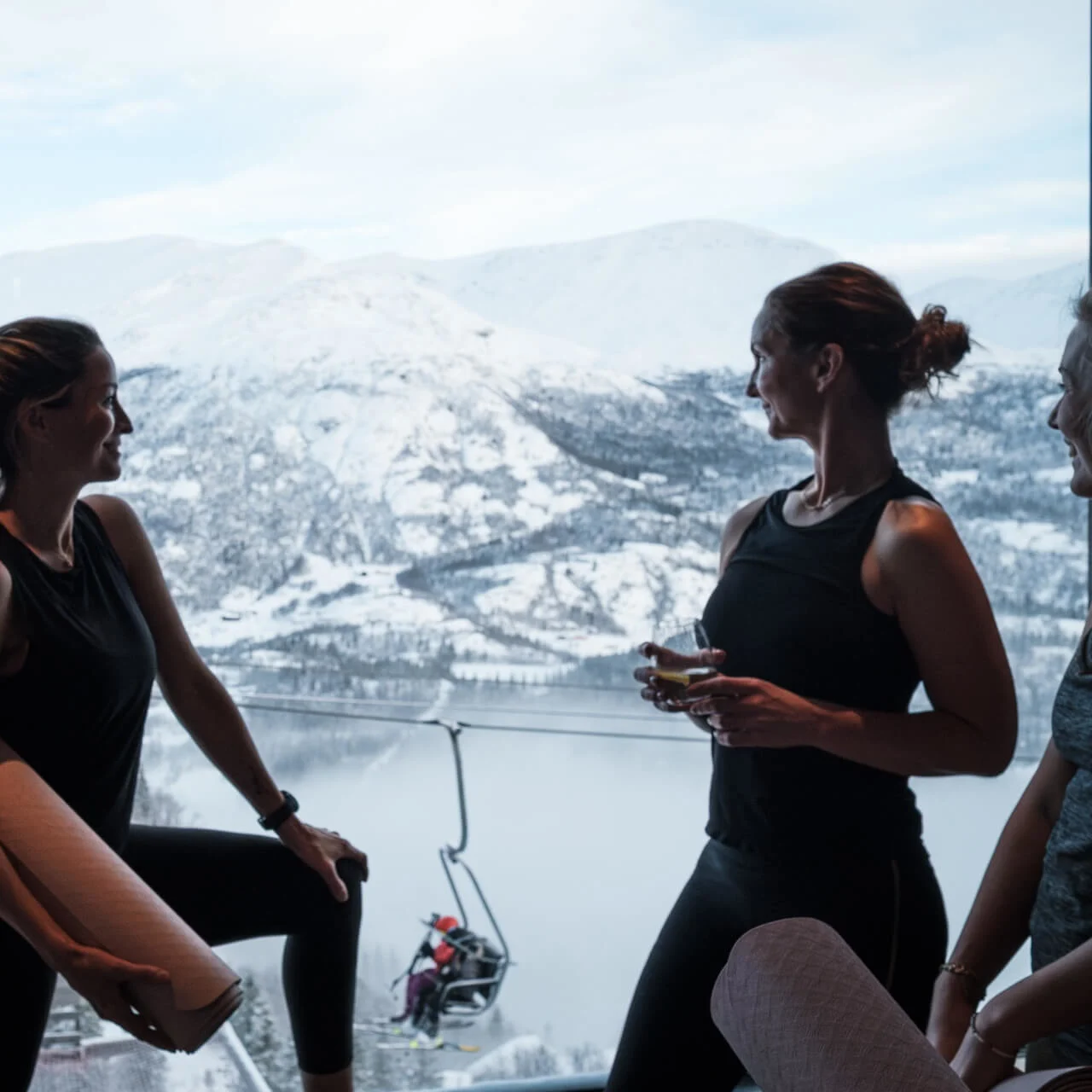 Four people, standing indoors, hold drinks and yoga mats while looking at snowy mountains through a large window. Ski lift and snowy landscape visible outside.