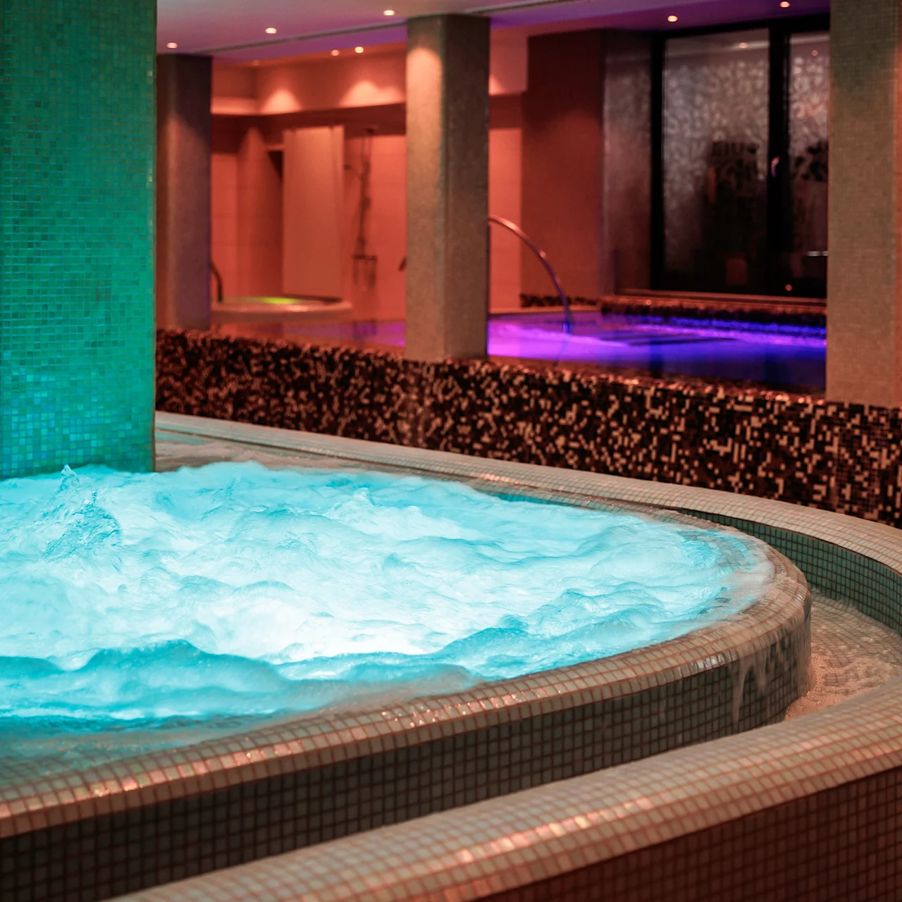 A large, illuminated Jacuzzi bubbles actively in a dimly lit spa area, surrounded by columns and decorative tiling, beside a reflective indoor pool.
