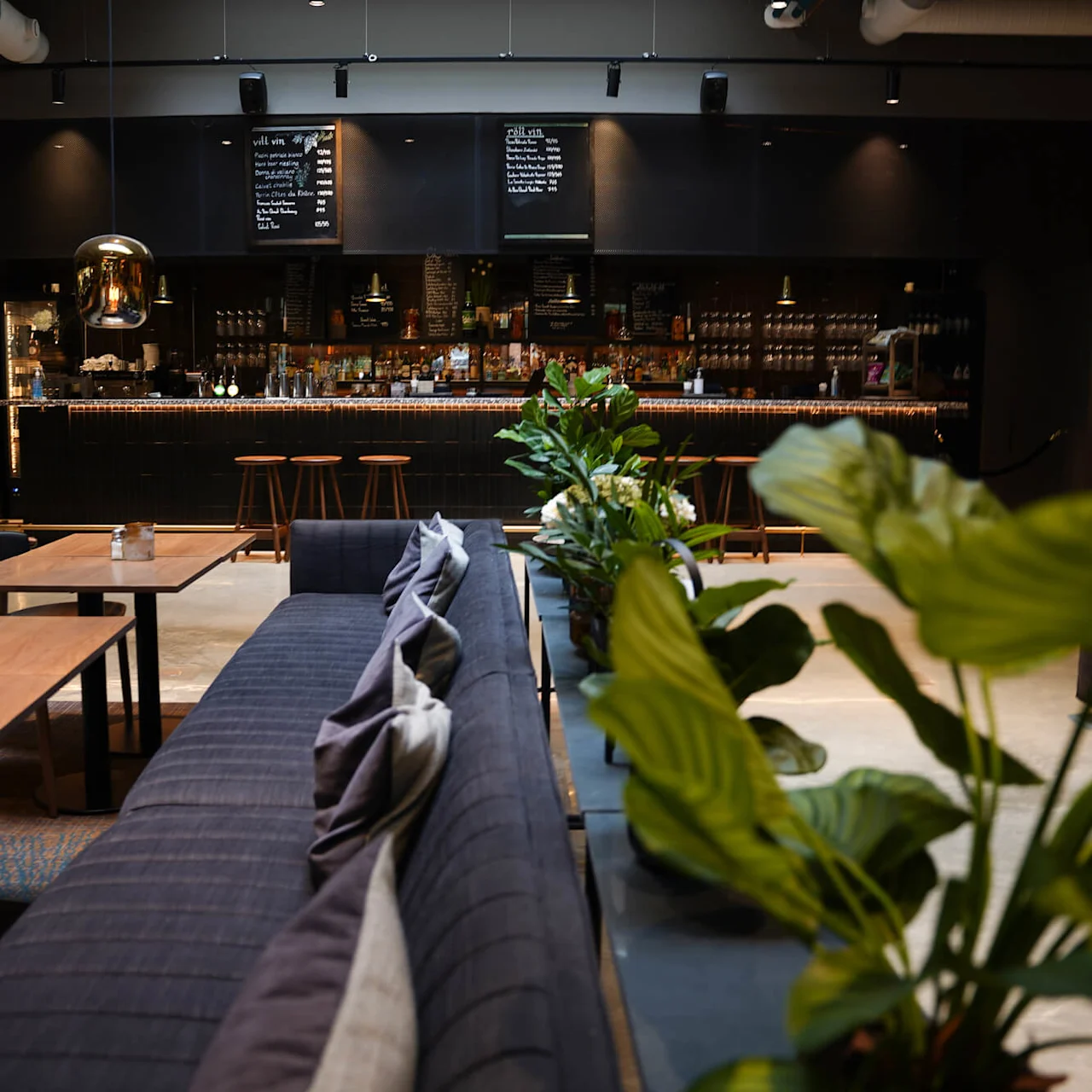 A cozy cafe interior with wooden tables, comfortable seating, pendant lights, and a bar with a menu board.