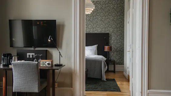 Et fladskærms-tv er monteret på en væg over et skrivebord og en stol, ved siden af en åben dørovergang, der afslører et soveværelse med seng, mønstret tapet og trægulve.