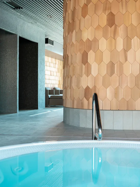 Badkar med metallhandtag står i ett elegant, modernt spa-interiör med sexkantiga träpaneler och grå kaklade väggar, under ett slattak med stämningsbelysning.
