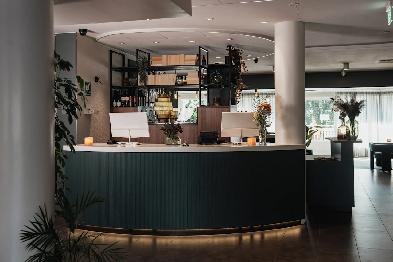 En elegant hotellobby med en mörkgrön receptionsdisk, datorer, blomsterarrangemang och en välfylld bar.