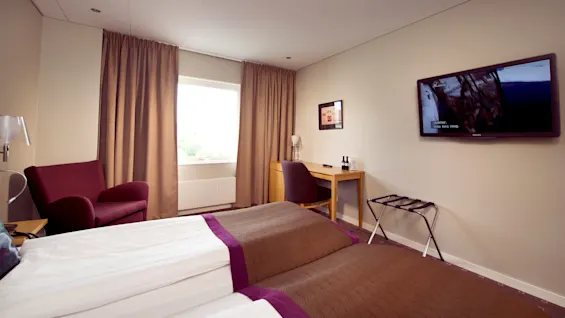A hotel room with a bed, armchair, desk, and mounted television displaying a scene with subtitles: "...skakar lite bra nog."