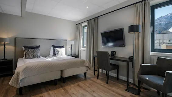 Sengen står sirligt redt, flankeret af to lamper, i et moderne hotelværelse. Et skrivebord har en kedel og kop under et vægmonteret TV, med bjerge synlige gennem vinduet.