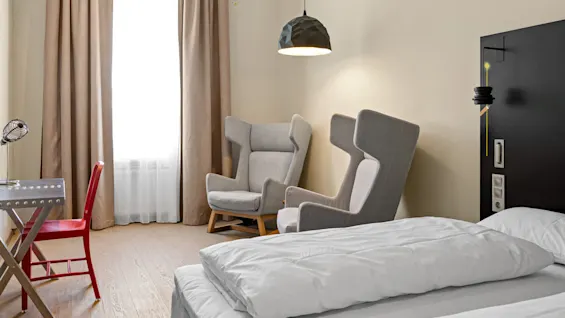 A bed with white linens is in a modern room. Gray armchairs sit beneath a pendant light, next to floor-length curtains. A desk with a red chair is nearby.