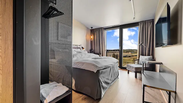 Et moderne hotellrom med en pent redd seng, vendt mot glassdører som leder til en utendørsutsikt. Tregulv og minimalistiske møbler forbedrer den rene, sofistikerte atmosfæren.