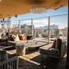 Tagterrasse med komfortable sofaer, et sofabord og udsigt over byen gennem store vinduer på en solrig dag.