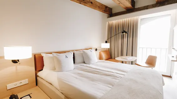 Ett ljust och modernt hotellrum med en stor säng, takbjälkar i trä och en liten sittgrupp vid ett fönster.