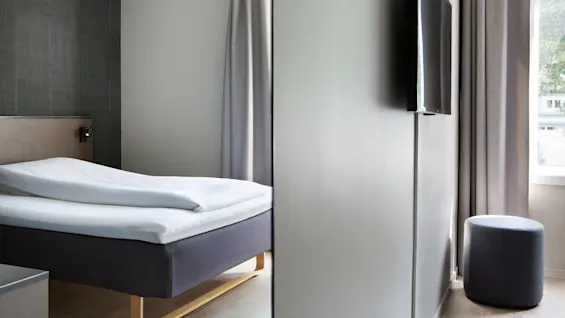 En pænt redt seng med en hvid dyne står i et moderne rum med en vægmonteret TV, grå gardiner og en cylindrisk skammel ved et vindue.