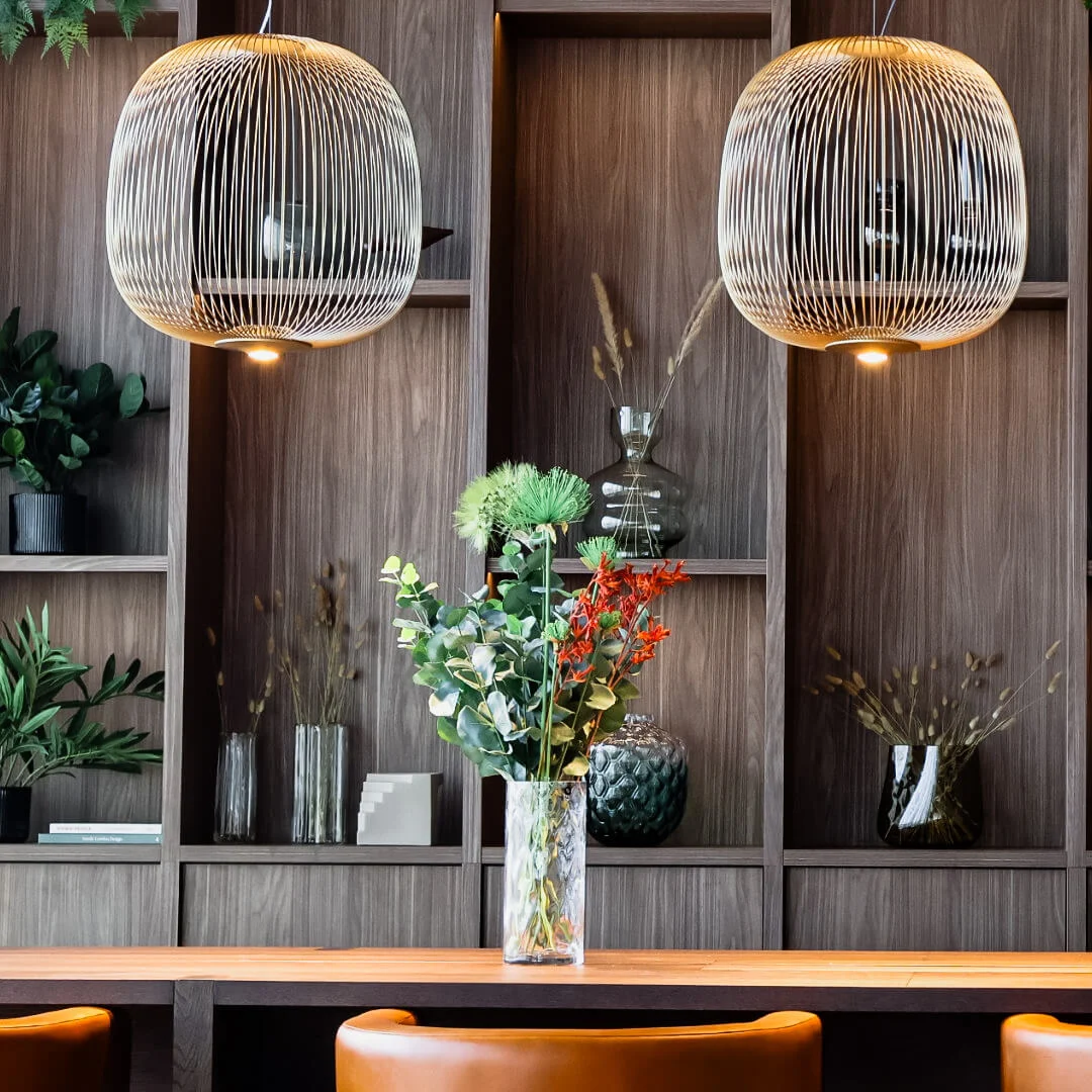 Suspended lights hang above a wooden table adorned with a vase of flowers. Wooden shelves in the background hold various decorative items and plants, creating a cozy atmosphere.