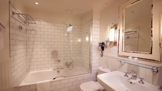Et badeværelse med badekar/brusekombination, toilet, vask og spejl, varmt belyst med et vintage æstetisk udtryk.