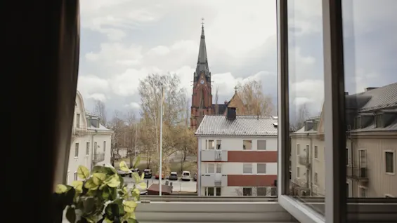 En kyrktorn står högt utanför ett öppet fönster, omgiven av träd och bostadshus. En krukväxt står på fönsterbrädan.