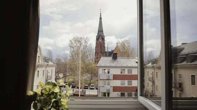 A church tower stands tall outside an open window, surrounded by trees and residential buildings. A potted plant rests on the windowsill.