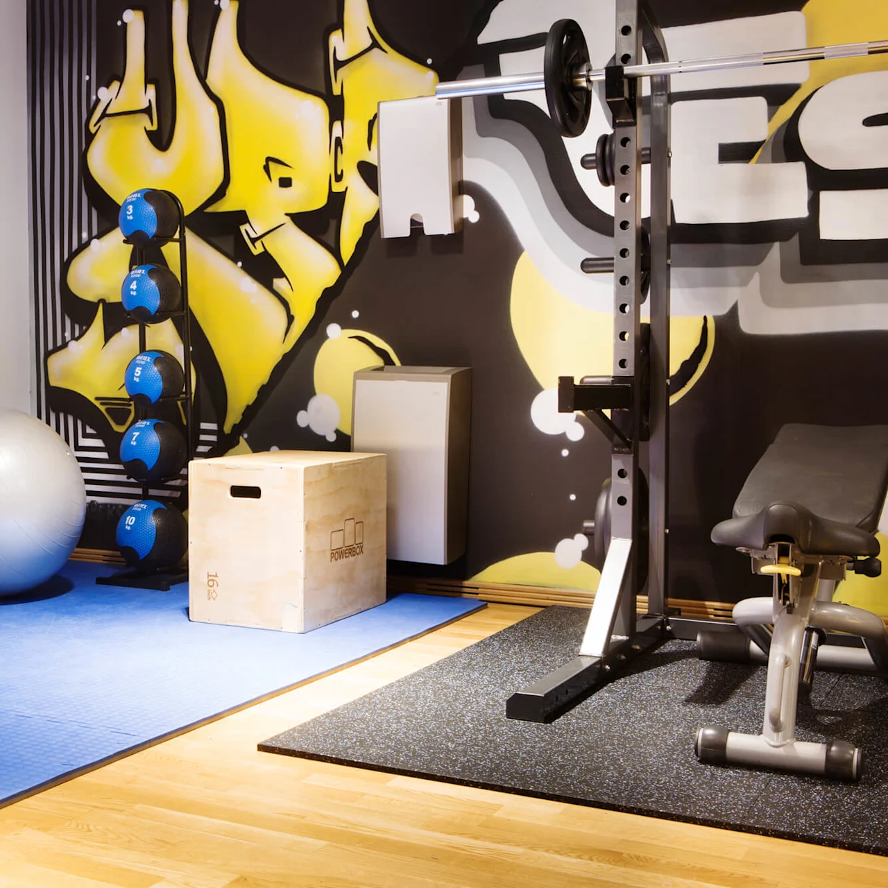 Exercise equipment, including kettlebells, resistance bands, a workout bench, and a fitness ball, are arranged in a gym. A colorful graffiti-style mural decorates the wall.