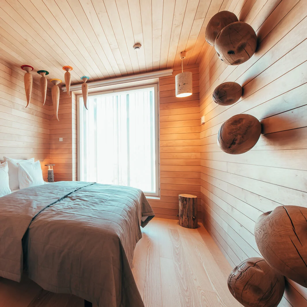 A wooden bed with white pillows rests in a cozy, warmly lit room featuring wooden paneling, unique ceiling sculptures, and wall-mounted round wood decor pieces beside a large window.