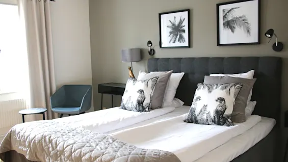 A cozy hotel room featuring a made double bed with decorative pillows, framed wall art, and an armchair by the window.