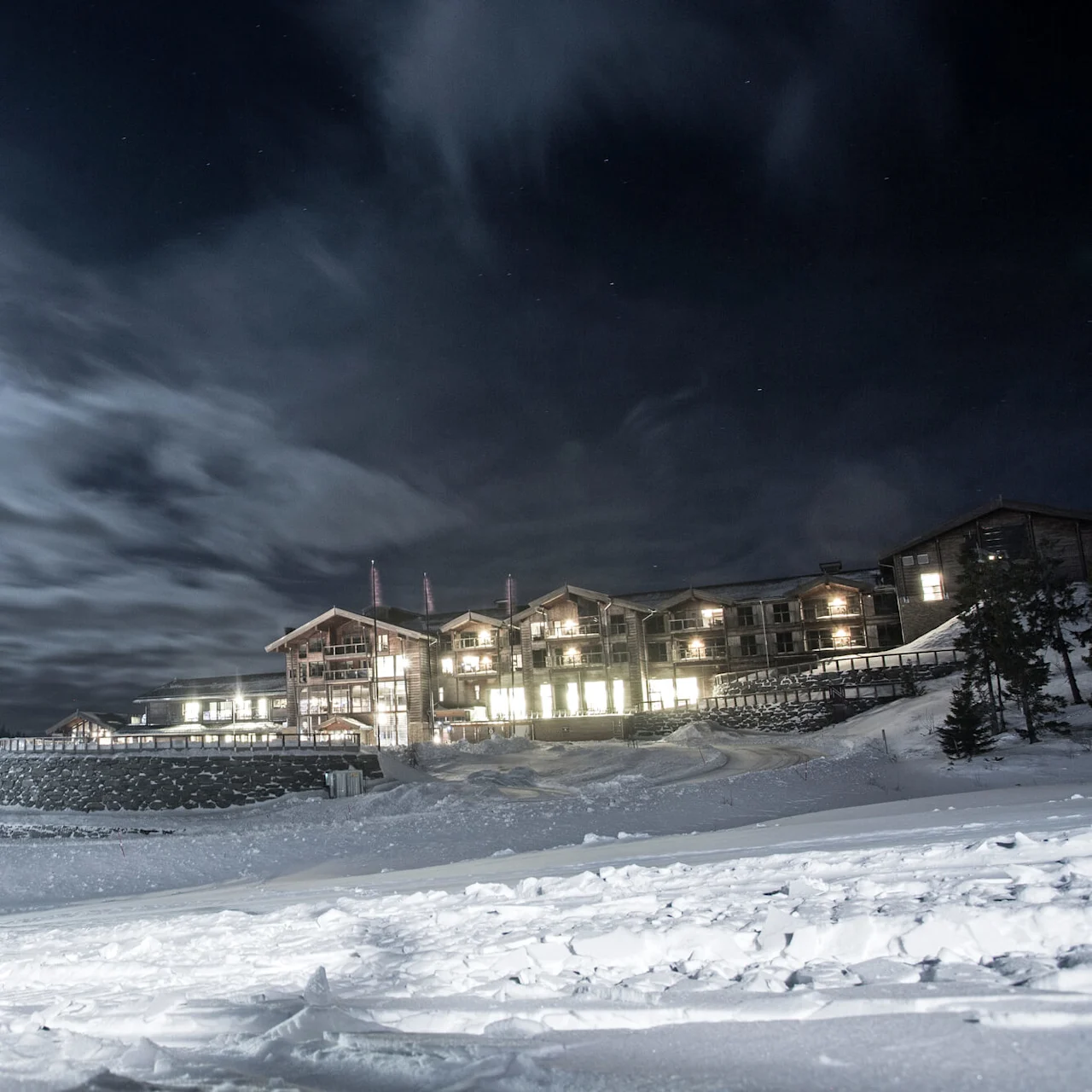 Snow-covered buildings glow with lights, surrounded by a wintry landscape under a bright, cloudy night sky with a visible full moon.