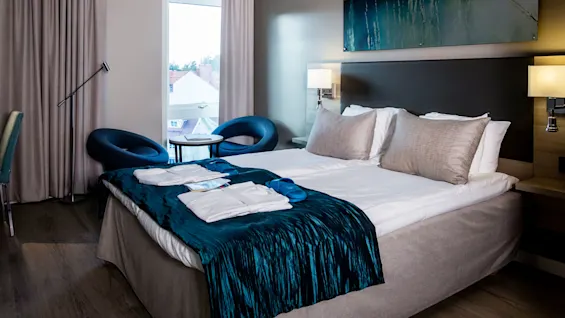 En pent hotelseng med et teal kast, badekåper og tøfler står i et moderne rom. Blå stoler og et lite bord ved vinduet har utsikt over andre bygninger.