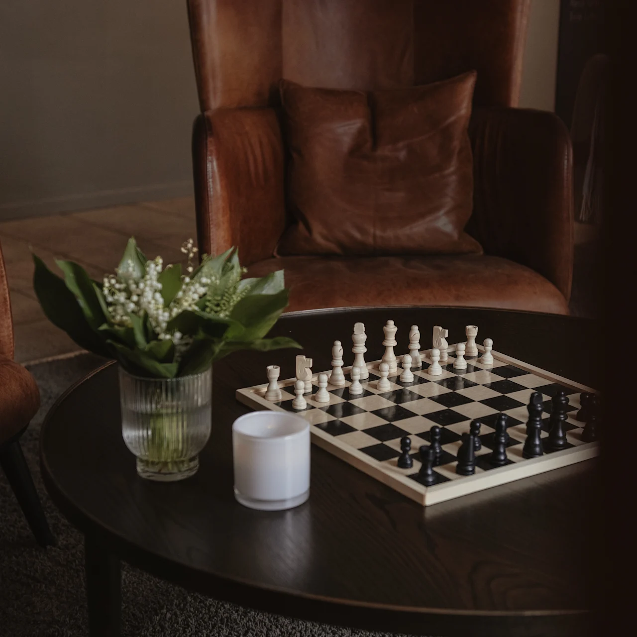 A chessboard with pieces set for play rests on a wooden coffee table, alongside a glass vase of white flowers, in a cozy room with leather armchairs.