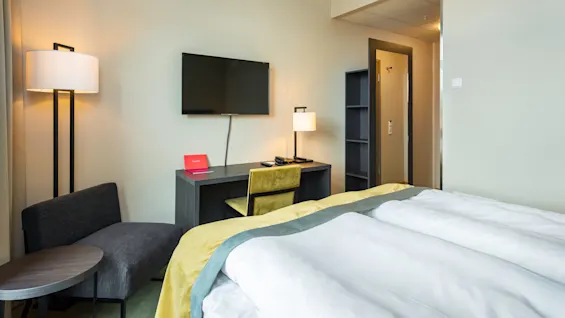 Moderni hotellihuone, jossa siististi pedattu sänky, kirjoituspöytä puhelimen ja muistivihkon kera, taulu-TV ja sohva, puhtaassa ja hyvin valaistussa tilassa.