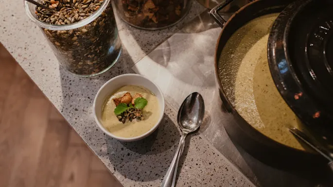 Kremet suppe i en bolle med krutonger og frø, ved siden av en stor gryte og toppingglass, som antyder et selvbetjent måltid.