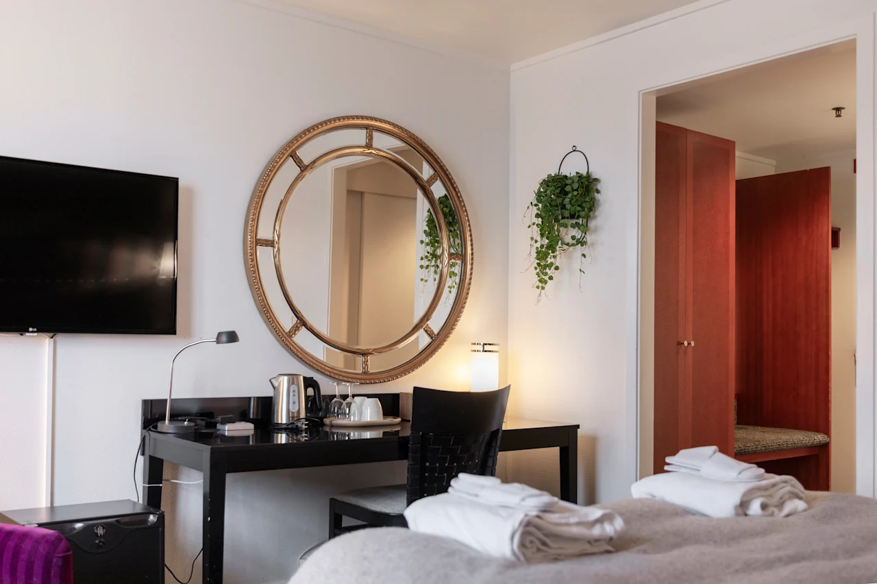 Viihtyisä hotellihuone, jossa on sänky puhtailla pyyhkeillä, työpöytä televisiolla ja koristeellinen pyöreä peili.