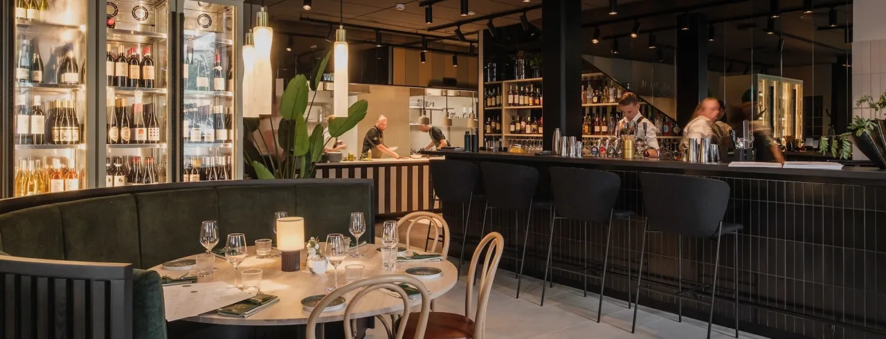 Elegant restauranginteriör med middagsgäster sittande vid bord och en bartender bakom en bar, upplyst av hängande lampor i en modern miljö.