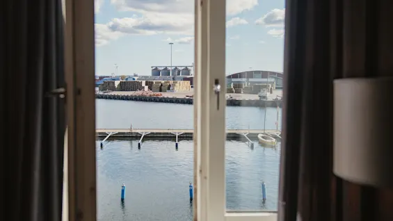 En naturskøn udsigt fra et vindue over en arbejdshavn med industribygninger, stablede varer og roligt vand. En bøje mærket 2 er synlig.