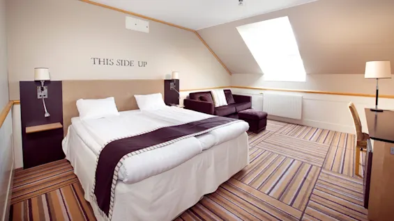 Ett välordnat hotellrum med dubbelsäng, soffa och skrivbord under snedtak. Text på väggen lyder "THIS SIDE UP".
