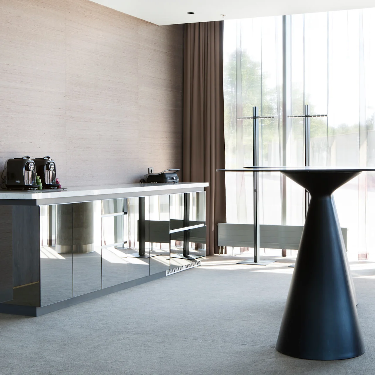 A modern, minimalistic room features a sleek black pedestal table near large windows, with a mirrored cabinet, coffee machines, and brown curtains, bathed in natural light.