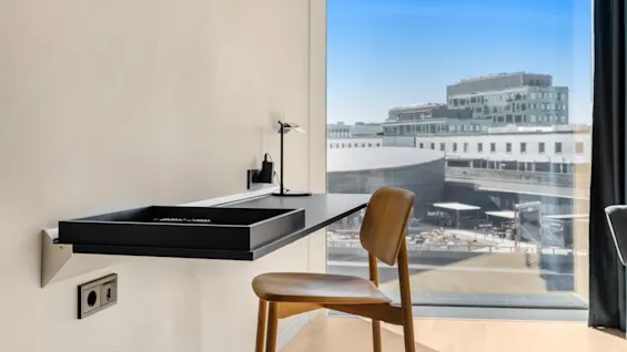 Moderne skrivebordsområde med træstol og lampe, med lys byudsigt gennem et stort vindue.