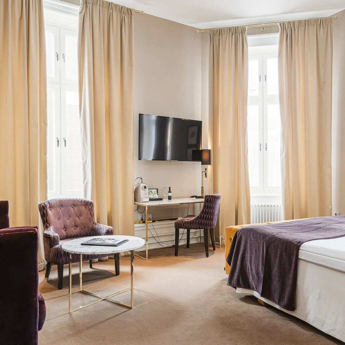 Elegant hotellrum med plyschsoffa, stol, skrivbord och säng i en neutral färgsättning.