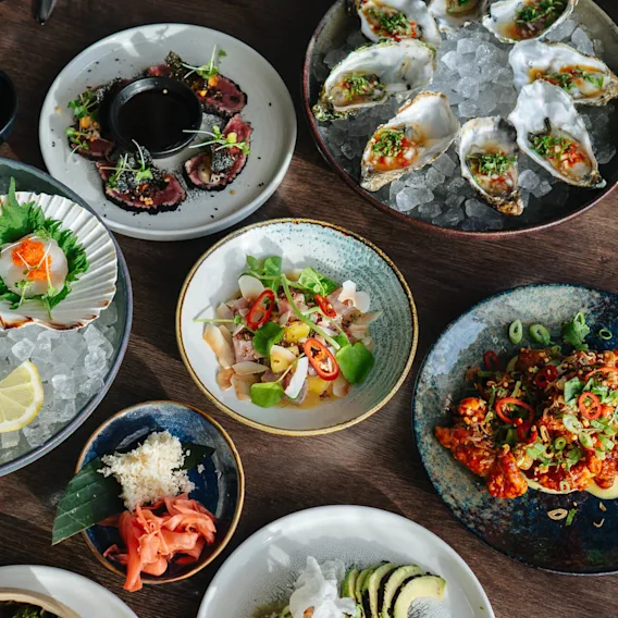 En mängd färgglada skaldjursrätter är konstnärligt arrangerade på ett träbord. Sashimi, ostron och ceviche pryds med örter, skivad chili och citronklyftor, omgivna av skålar och tallrikar.