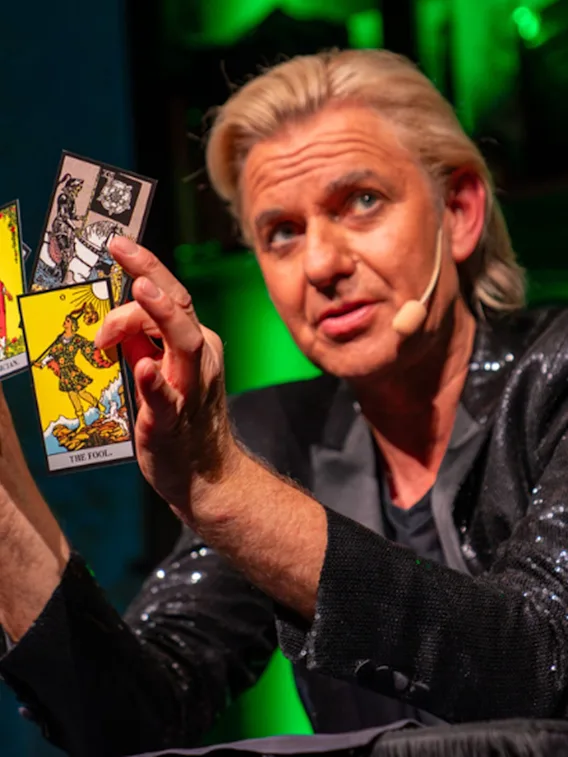 En person utför ett trolleritrick eller en tarotläsning, håller flera kort utspridda i handen på en grönbelyst scen.