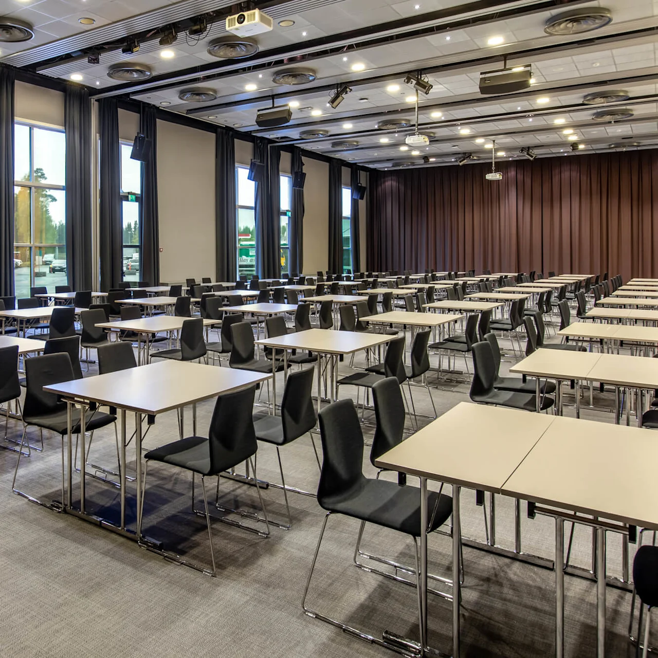 Empty conference room with rows of black chairs and beige tables. Windows line the side wall, with curtains partially drawn and lights on the ceiling. An exit sign indicates the door.