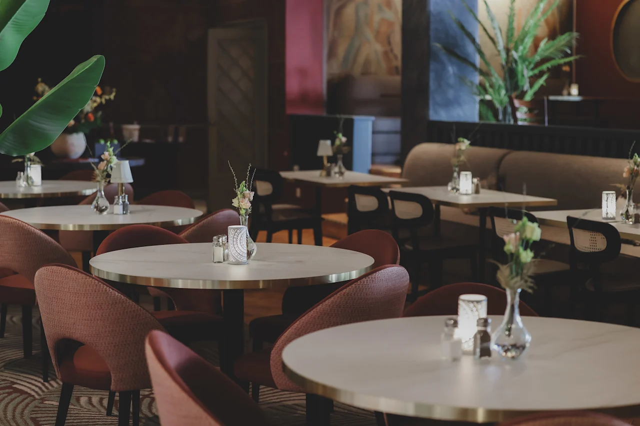 Tables are set with small vases and candles in a dimly lit, elegant restaurant with red chairs and potted plants. The decor includes artwork on the walls, creating a sophisticated atmosphere.