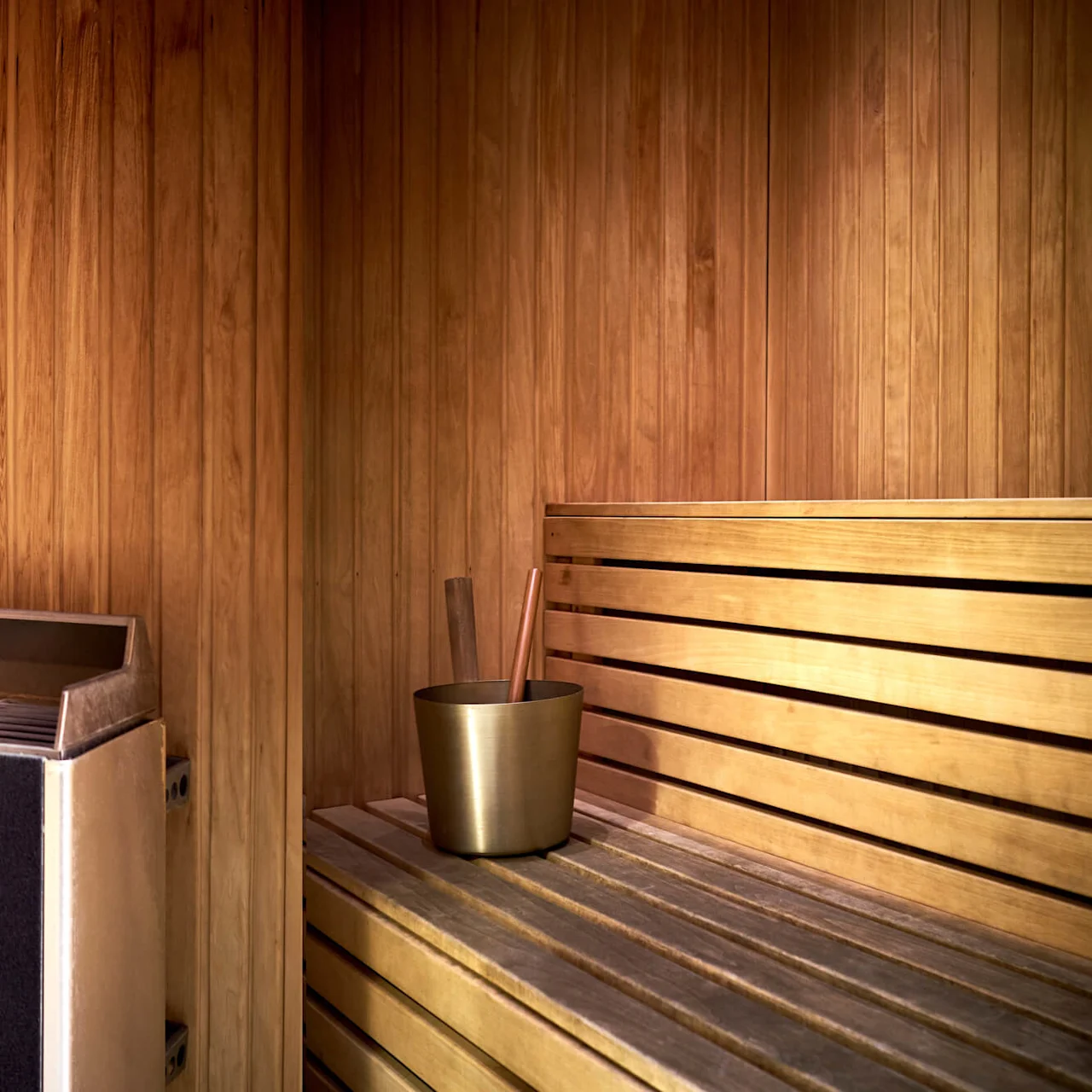 A sauna interior with wooden benches and walls, a heater filled with stones, and a metal bucket with a ladle.