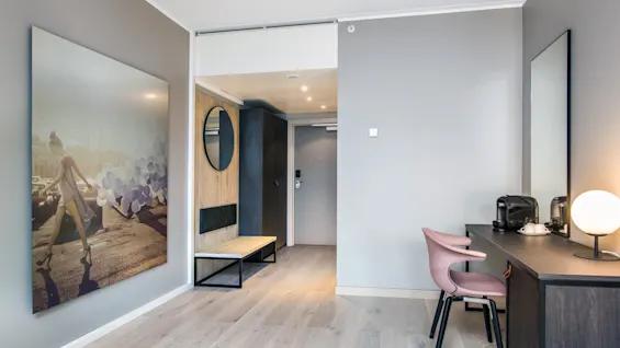 A hotel room with modern decor features a desk with a coffee maker and lamp. A large photo of a woman with balloons is on the wall beside the entrance.