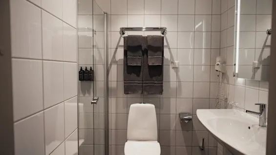 Ett ljust och funktionellt hotellbadrum med vitkaklade väggar, en toalett, en glasdusch och ett väl upplyst handfat.
