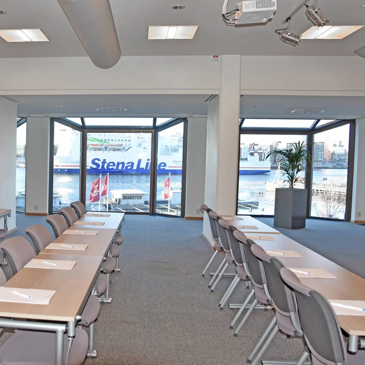 Meeting room with arranged tables and chairs, overlooking a harbor through large windows. Text on the ship reads "Stena Line." Plants adorn the space, and natural light fills the room.