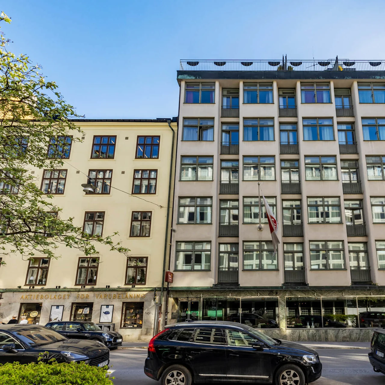 Ett modernt hus med stora fönster och en takterrass i solljus, beläget vid en stadsgata med parkerade bilar framför.