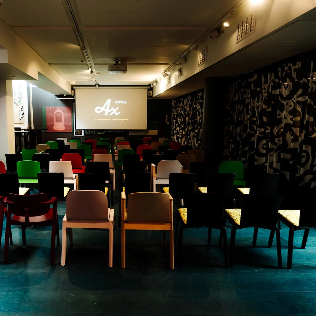 Rows of colorful chairs face a projector screen displaying "Hotel Ax" in a dimly lit room with artistic decor and paintings on the walls.