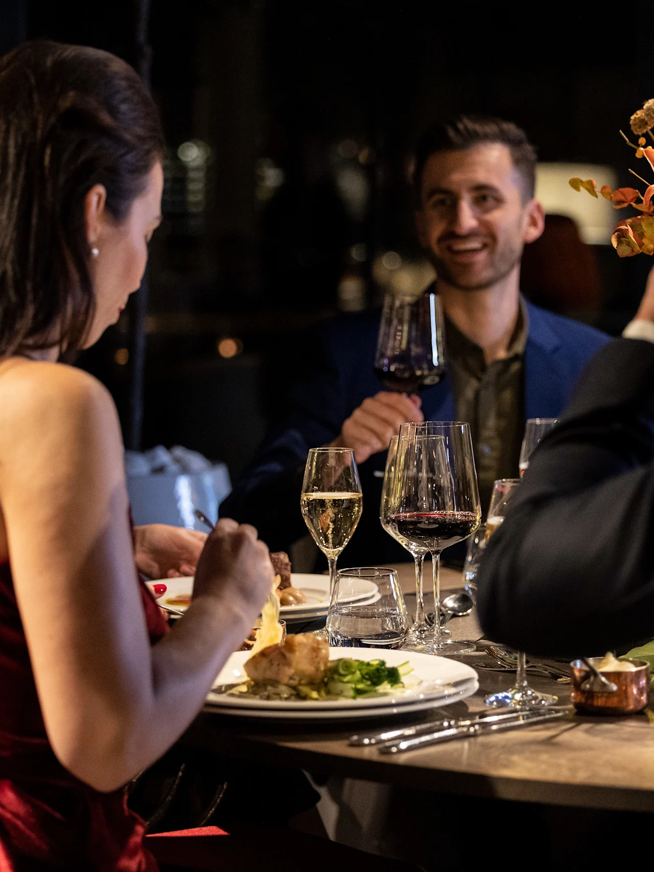 Norwegian Bokmål: Folk spiser ved et elegant dekket bord, deler smil og samtale. Vinglass og en blomsterdekorasjon forsterker den intime kveldssituasjonen.
